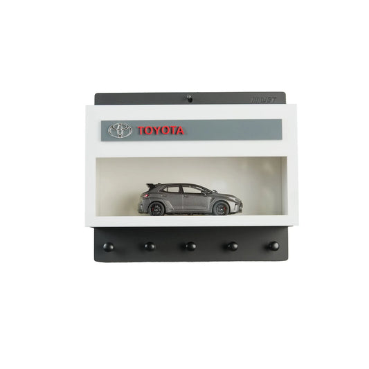 Black and white Toyota wall key hook rack with a gray scale model car inside a display case and five hooks below.