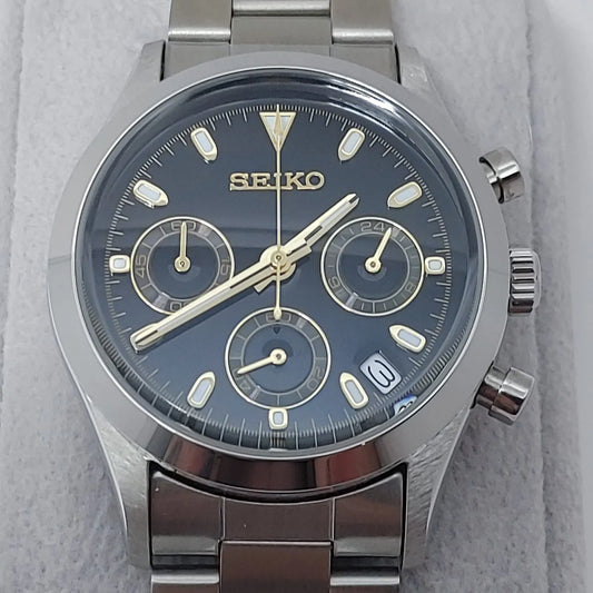 Seiko VK63 chronograph watch with black dial, gold-tone markers, three subdials, date window, and stainless steel bracelet.