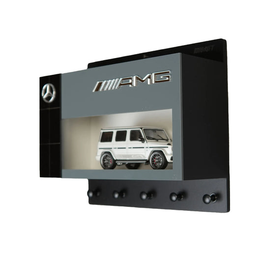 Black Mercedes AMG wall-mounted key hook rack with a white scale model G-Class car inside a display case.