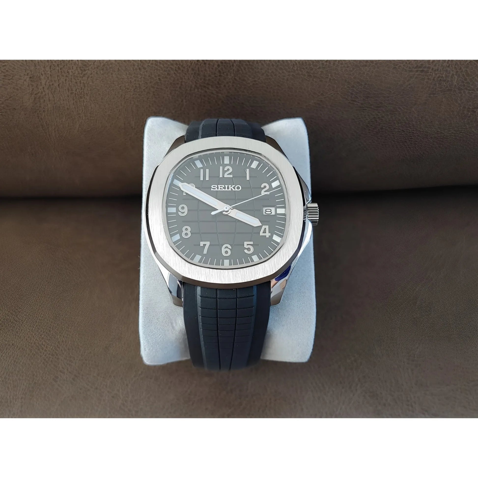 Seiko Aquanaut watch with black textured dial, silver case, white numerals, and black rubber strap on white cushion.