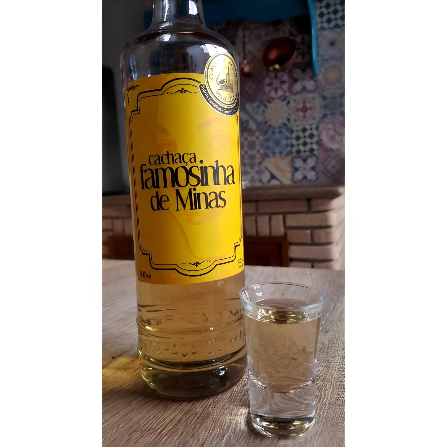 Tall clear glass bottle with yellow label reading "Cachaca Famosinha de Minas" next to a filled shot glass.