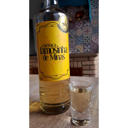 Tall clear glass bottle with yellow label reading "Cachaca Famosinha de Minas" next to a filled shot glass.