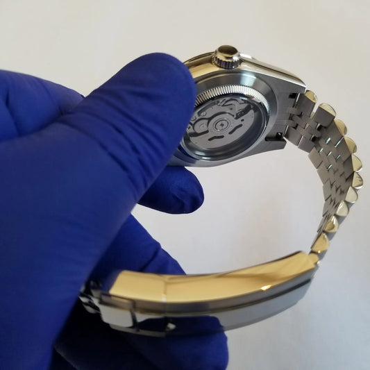 Silver stainless steel watch with transparent back, revealing automatic movement, held by hand in blue glove.