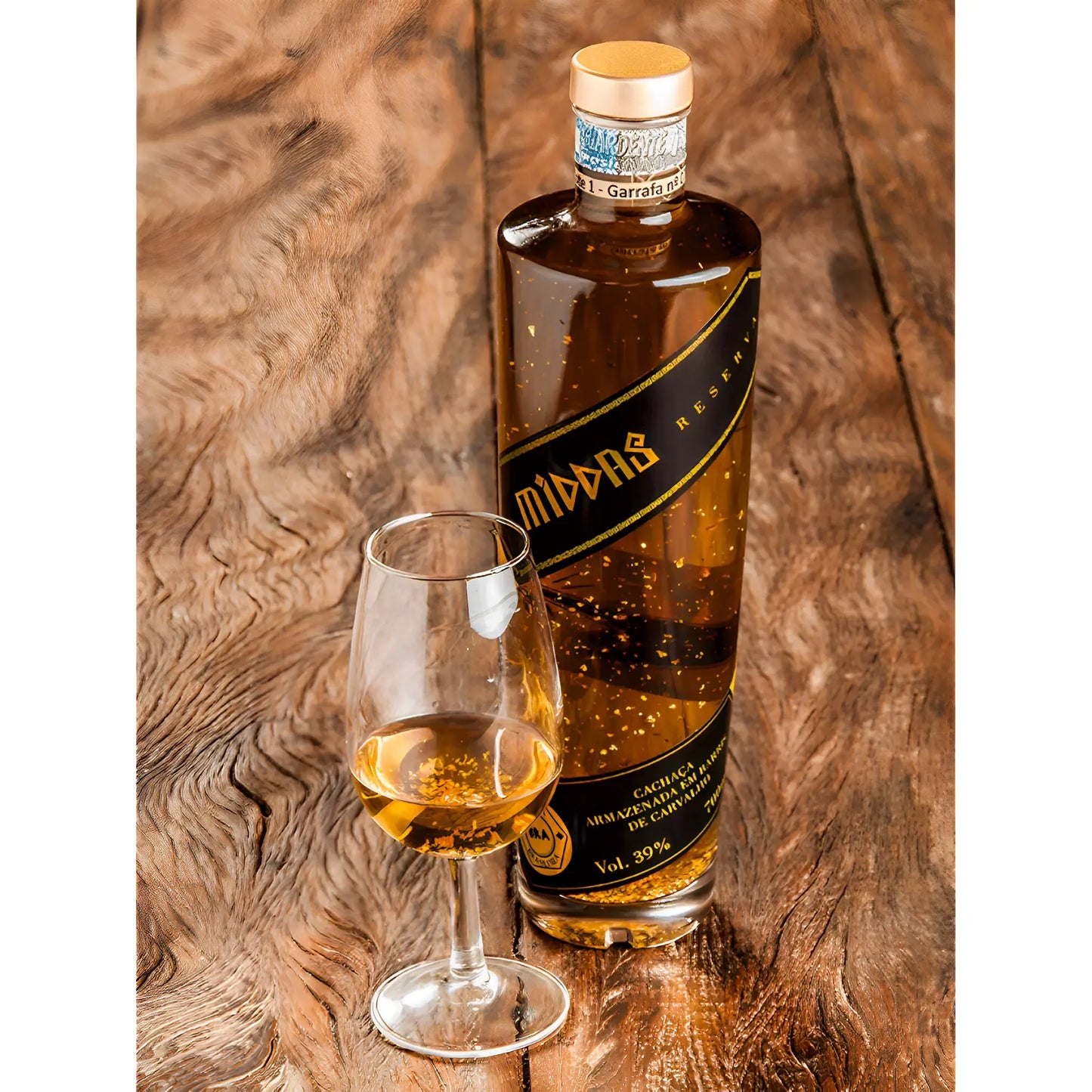 Tall clear bottle of golden cachaca with suspended 23k gold flakes and black label, beside a filled tasting glass on wooden.