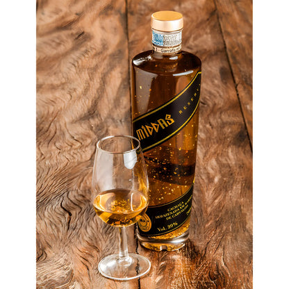 Tall clear bottle of golden cachaca with suspended 23k gold flakes and black label, beside a filled tasting glass on wooden.