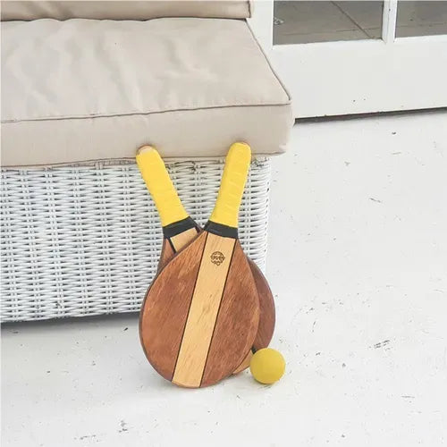 Wooden frescobol paddle racket with yellow handles and matching yellow ball on white floor next to wicker sofa.