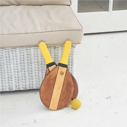 Wooden frescobol paddle racket with yellow handles and matching yellow ball on white floor next to wicker sofa.