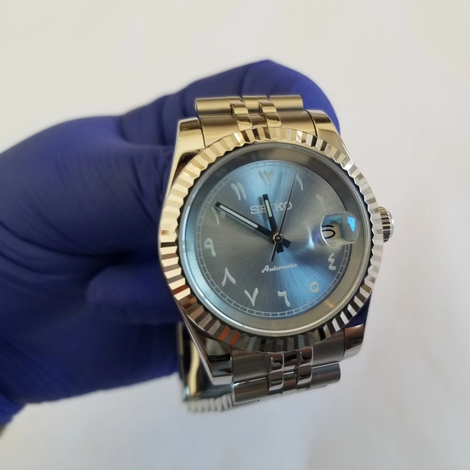 39mm silver stainless steel watch with fluted bezel, ice blue dial, white Arabic numerals, and date window at 3 o'clock.