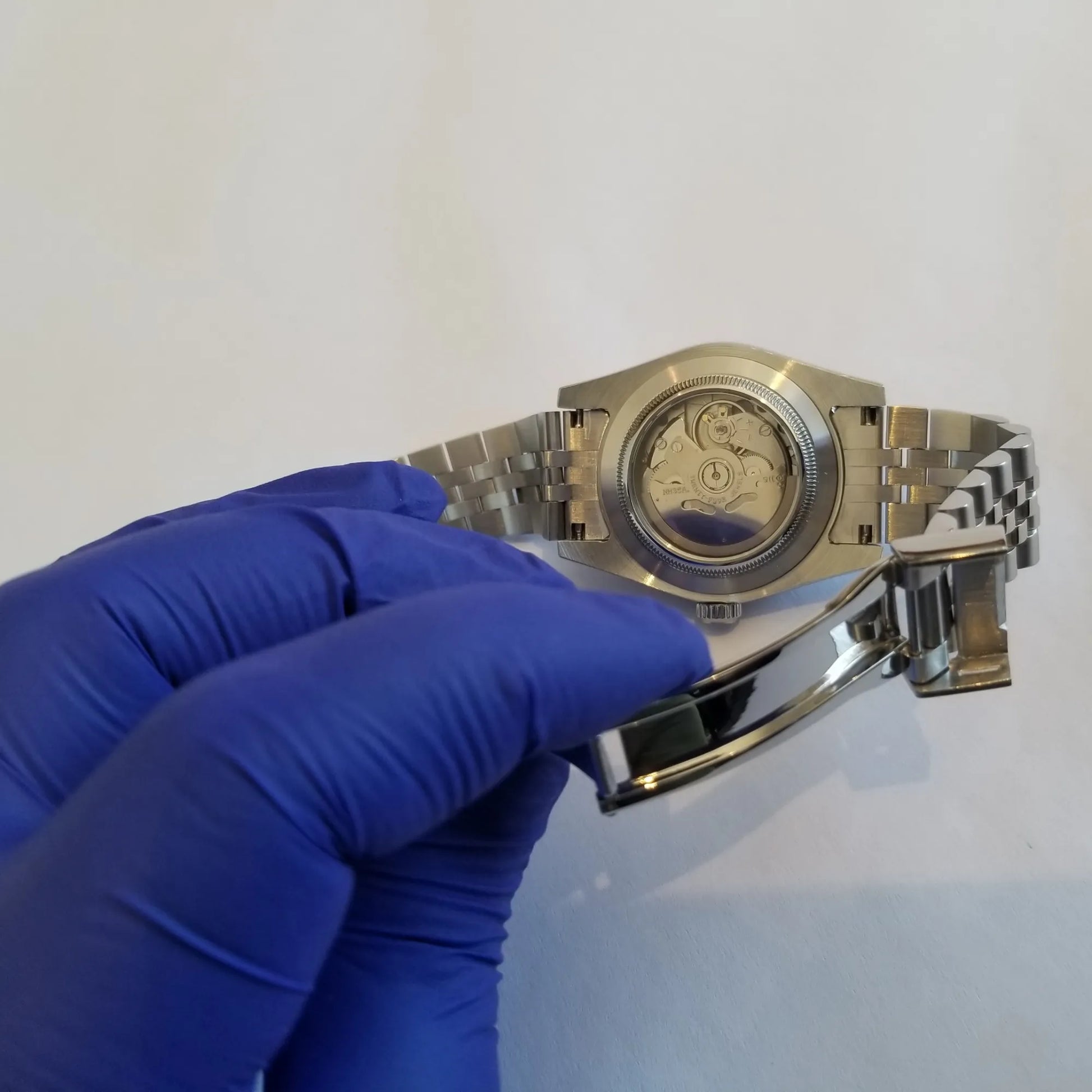 Silver stainless steel watch with fluted bezel and transparent case back showing movement, held by a hand in blue glove.