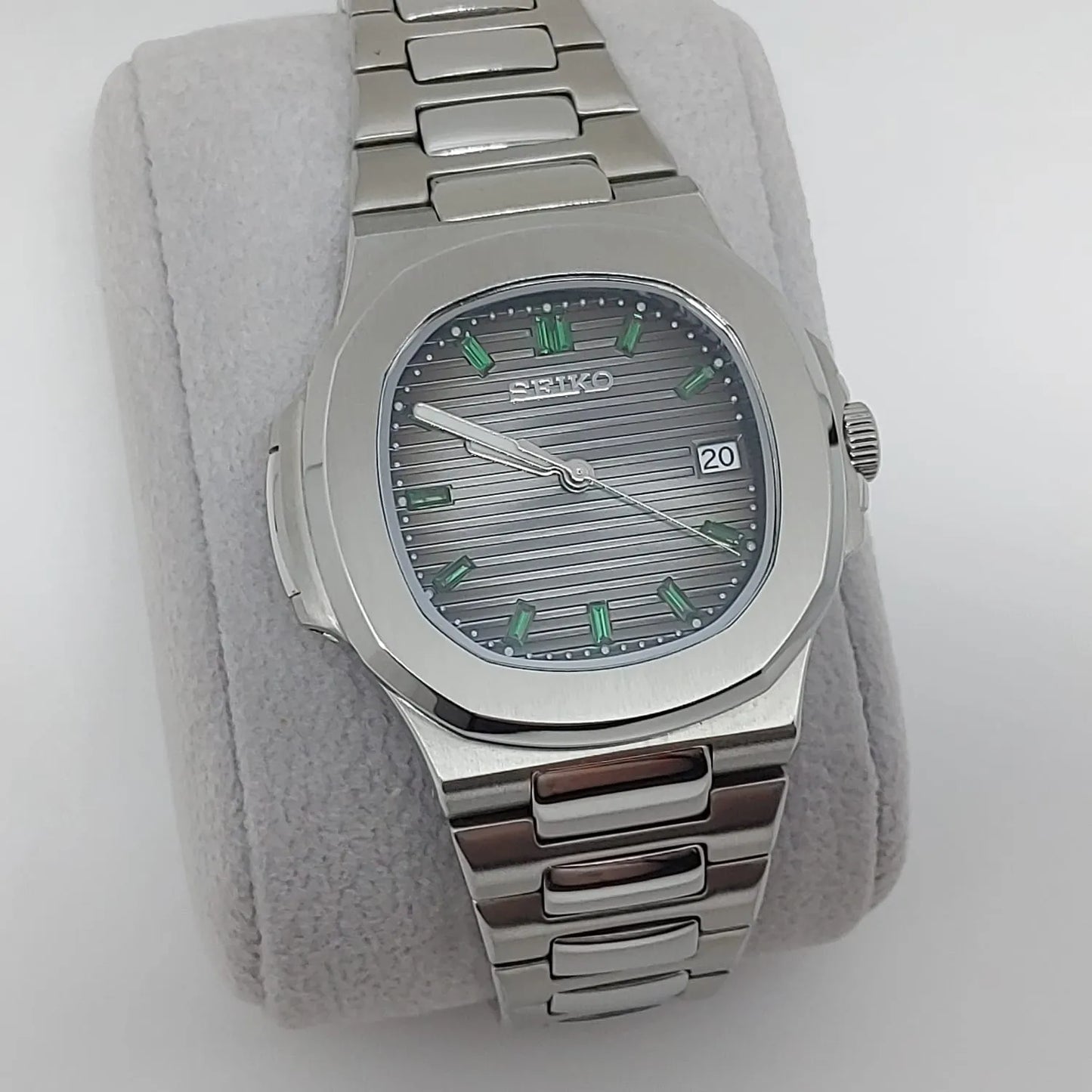 Seiko stainless steel watch with emerald green baguette markers, horizontal textured dial, and date window at 3 o'clock.