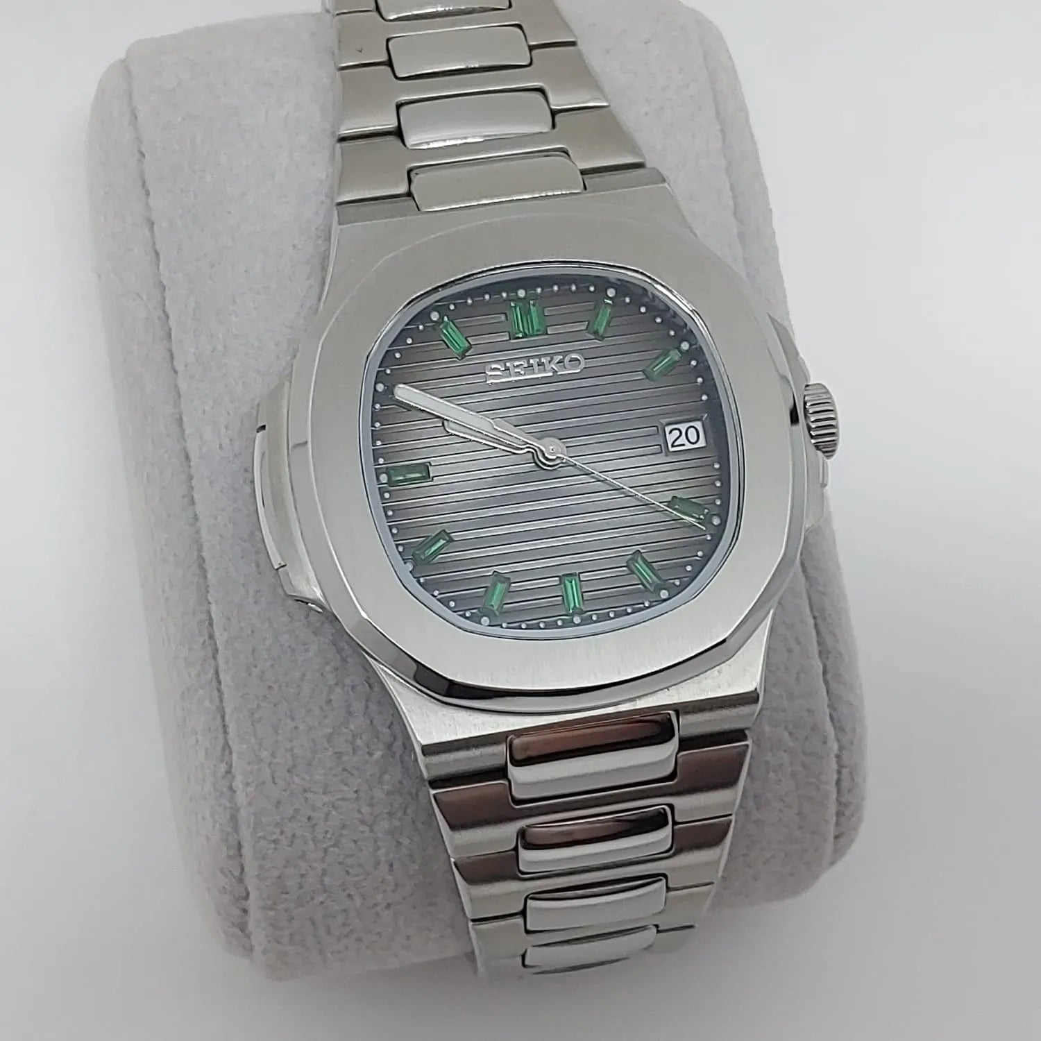 Seiko stainless steel watch with emerald green baguette markers, horizontal textured dial, and date window at 3 o'clock.