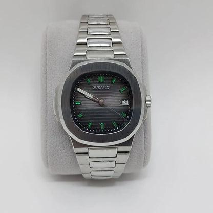 Stainless steel watch with rounded square bezel, black textured dial, green baguette hour markers, and date window at 3.
