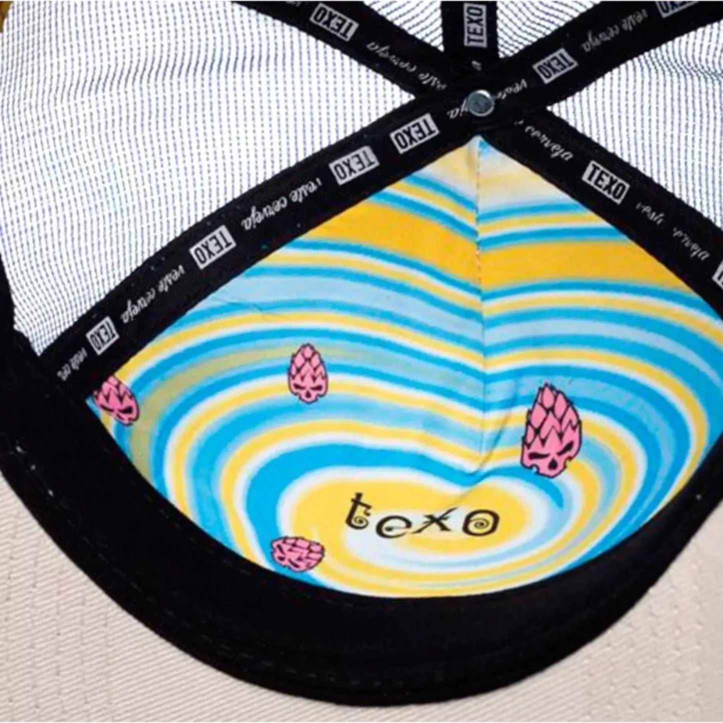 Inner view of hat with black lining, white mesh crown, and colorful yellow and blue swirl pattern featuring pink hop cone.