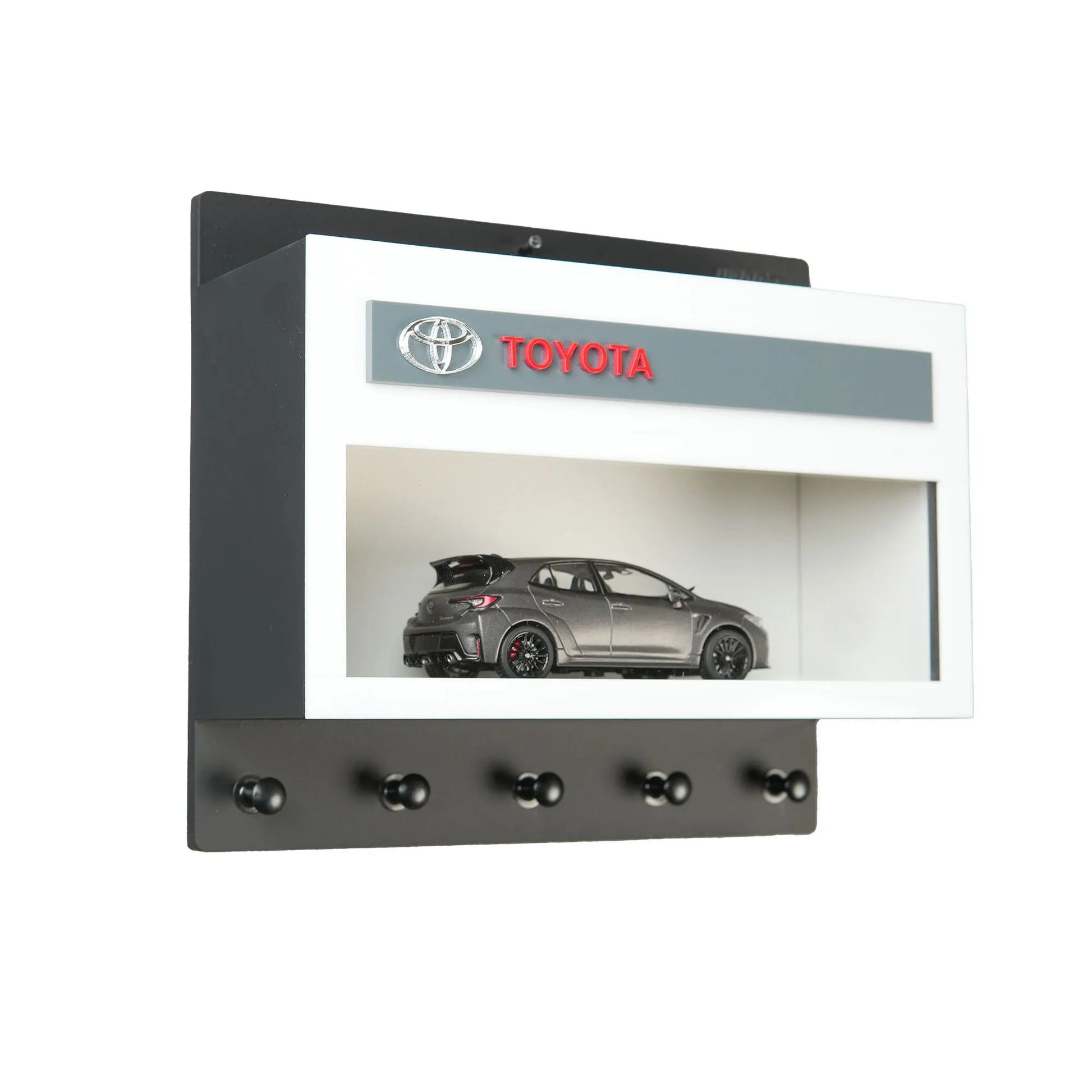 Black and white wall-mounted key hook rack with a Toyota logo and a gray scale model car displayed inside a glass case.