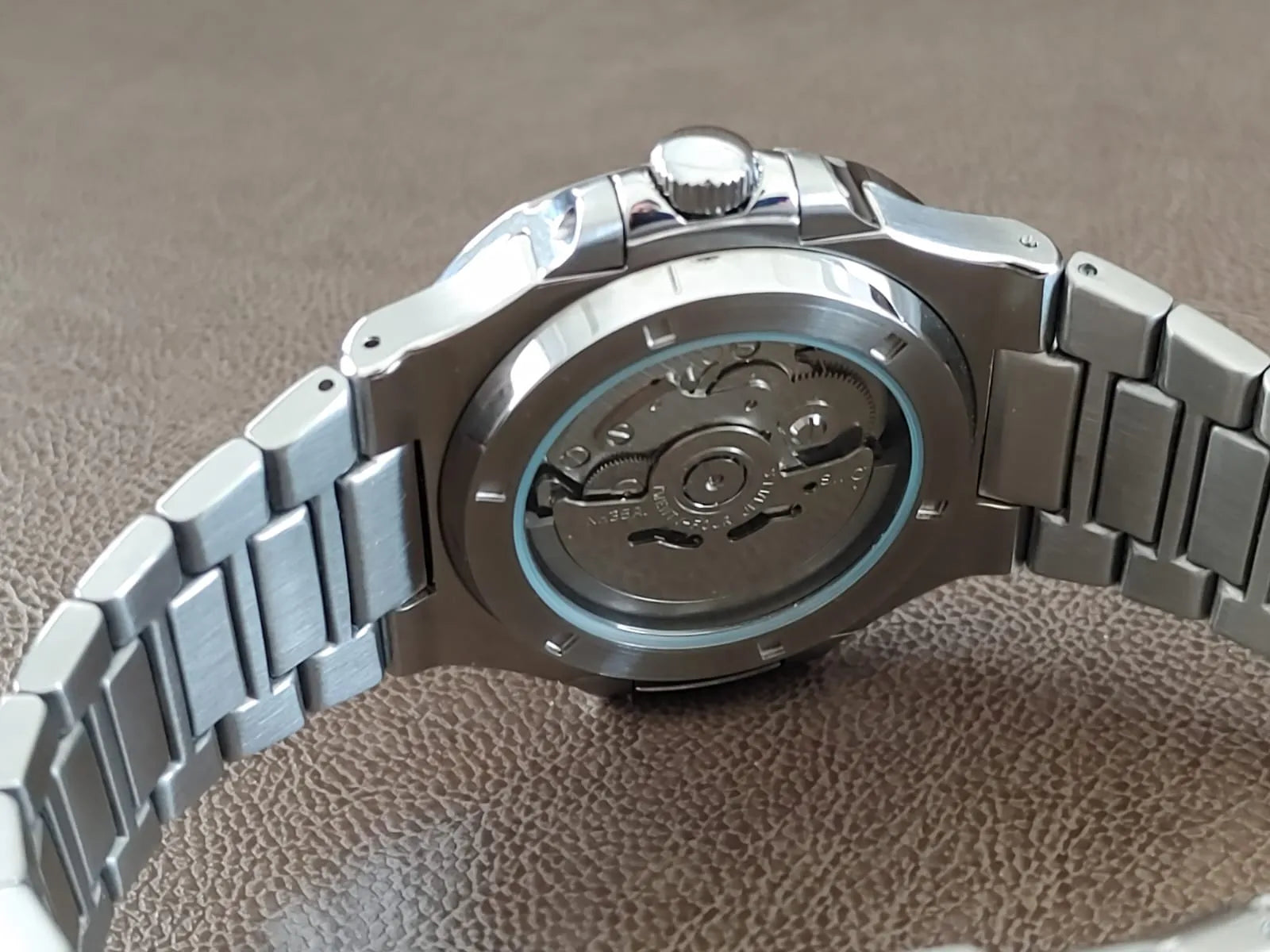 Stainless steel watch with visible automatic movement through transparent back case and linked metal bracelet.
