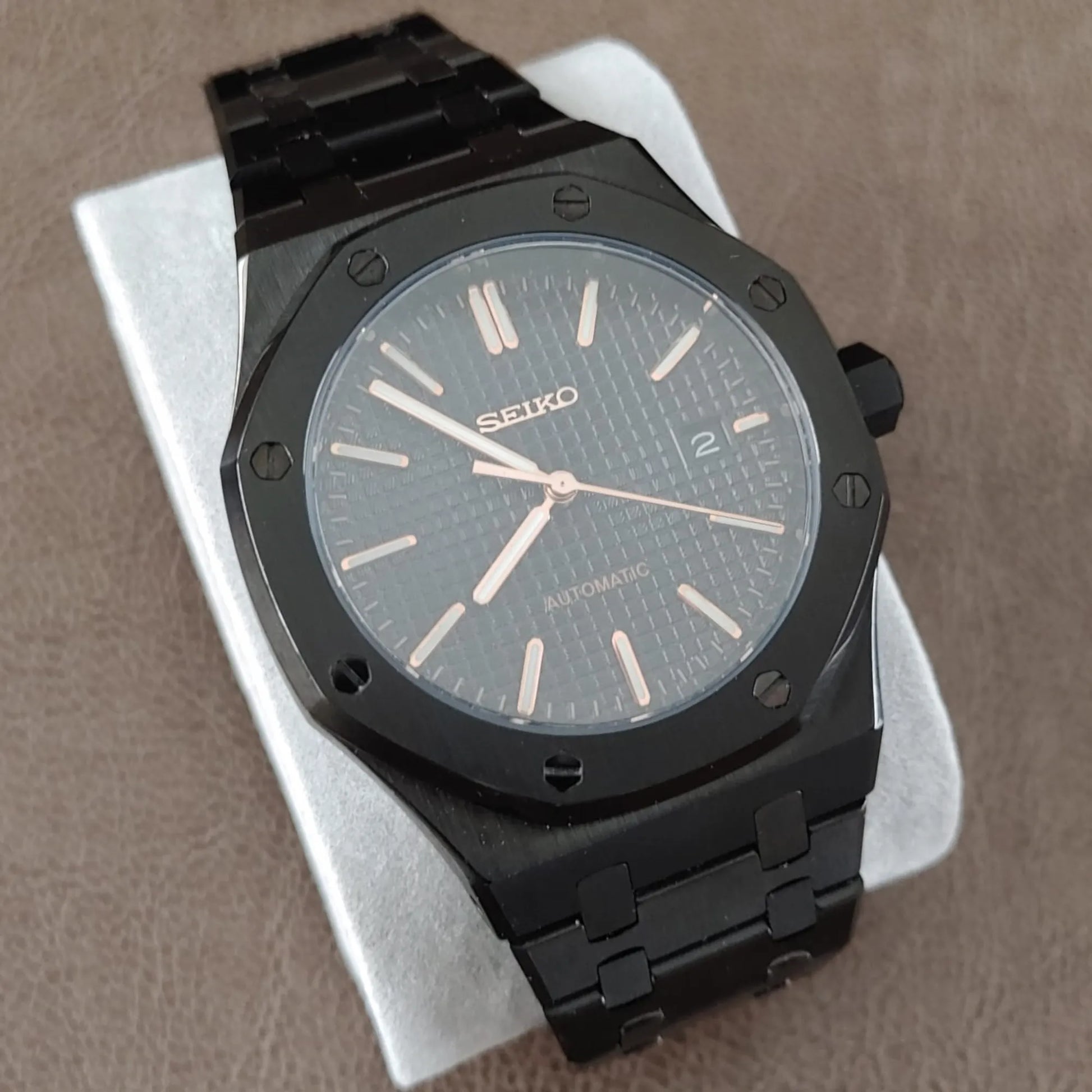 Black stainless steel Seiko watch with rose gold hour markers and hands, textured dial, and date window at 3 o'clock.