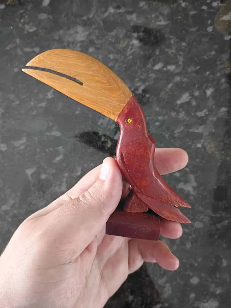 Handcrafted wooden toucan sculpture with natural wood and red-stained details held in hand against dark stone background.
