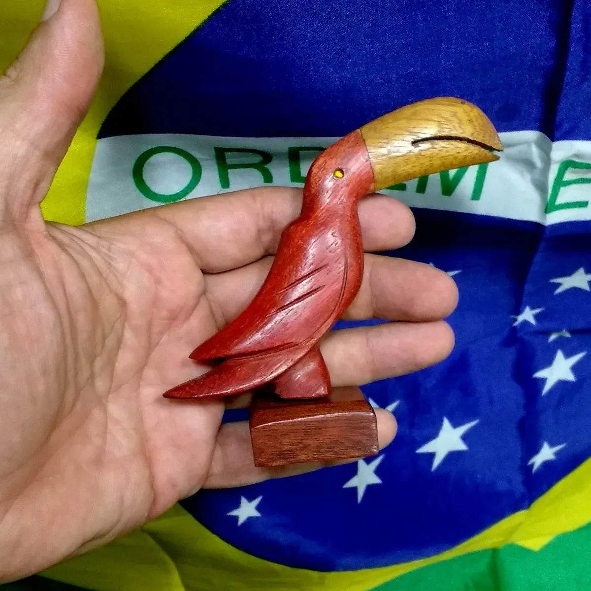Small handcrafted wooden toucan with a yellow beak and red body, held in hand against a Brazilian flag background.
