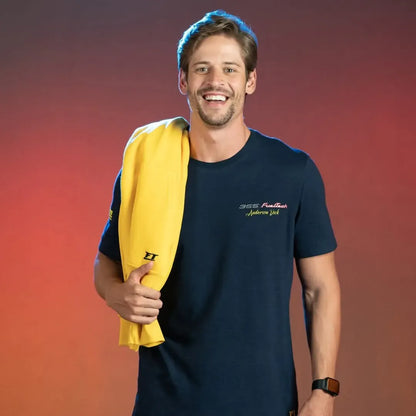 Navy blue short-sleeve t-shirt with small yellow text on left chest, worn by smiling man holding yellow sweatshirt over.