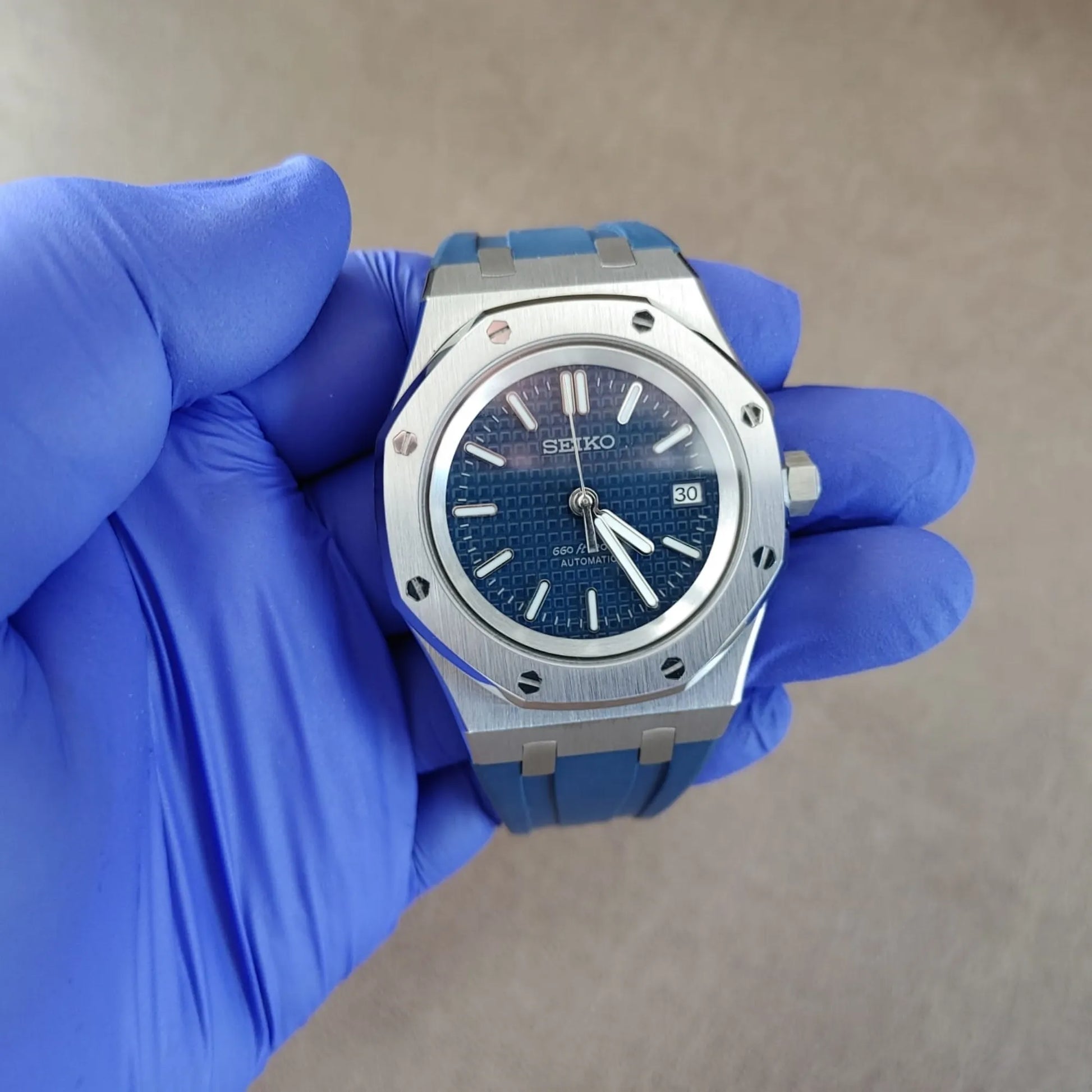 Seiko NH35 wristwatch with navy blue dial, silver octagonal bezel, and blue rubber strap held by gloved hand.