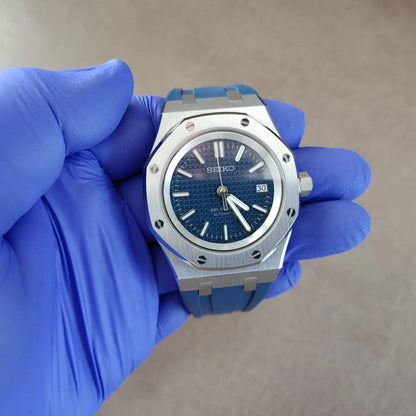 Seiko NH35 wristwatch with navy blue dial, silver octagonal bezel, and blue rubber strap held by gloved hand.