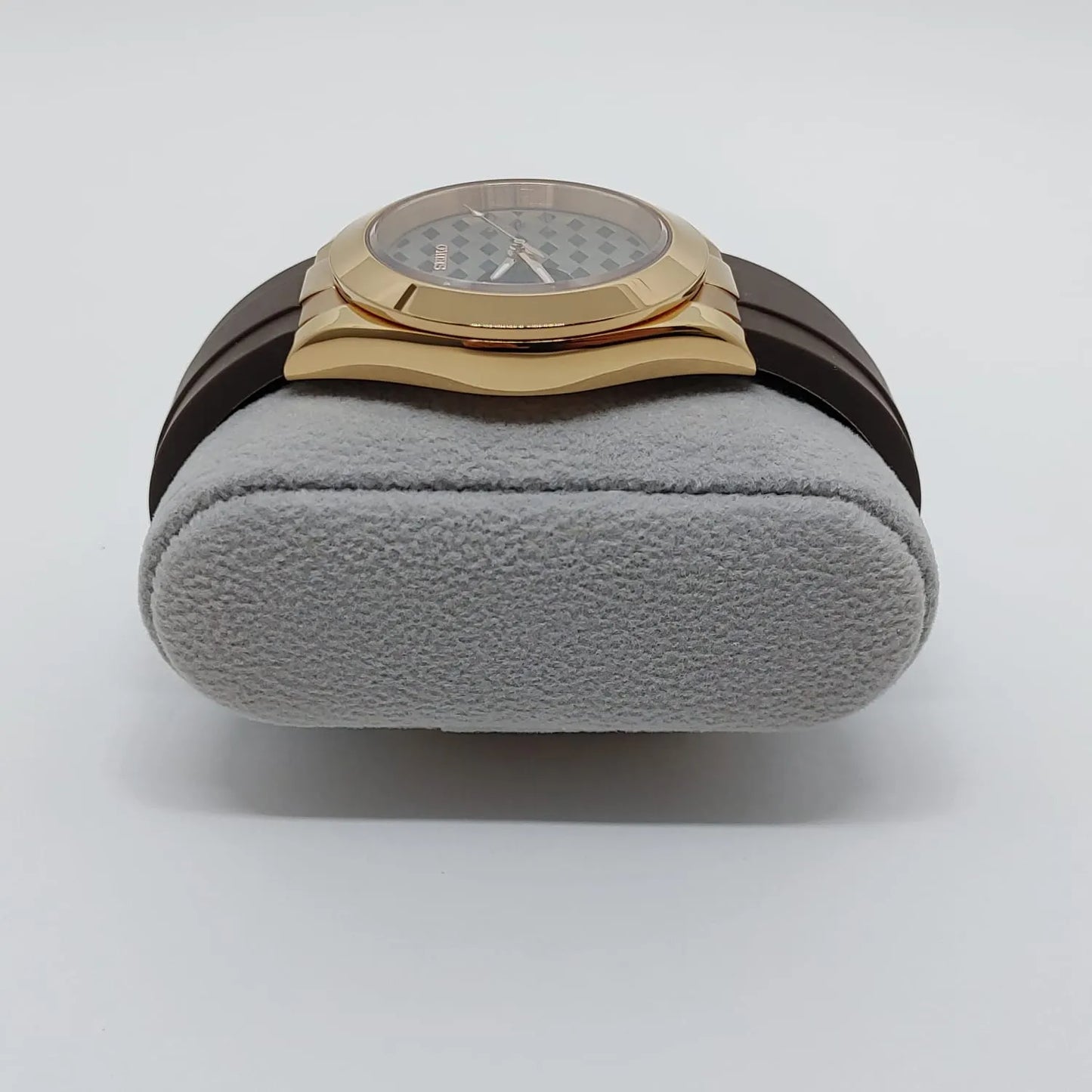 Gold-toned square watch case with rounded edges, brown leather strap, textured dial with guilloche pattern on gray cushion.