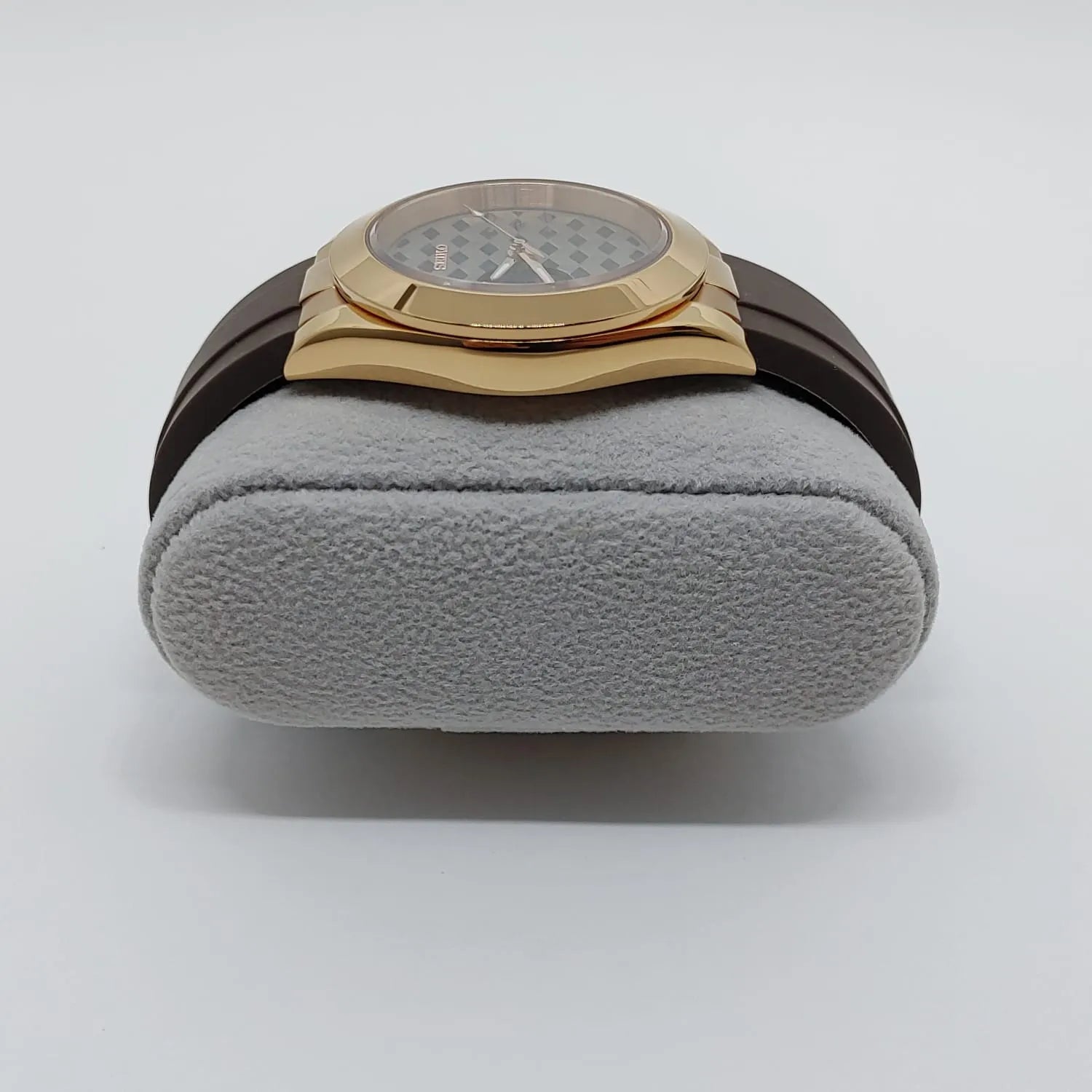 Gold-toned square watch case with rounded edges, brown leather strap, textured dial with guilloche pattern on gray cushion.