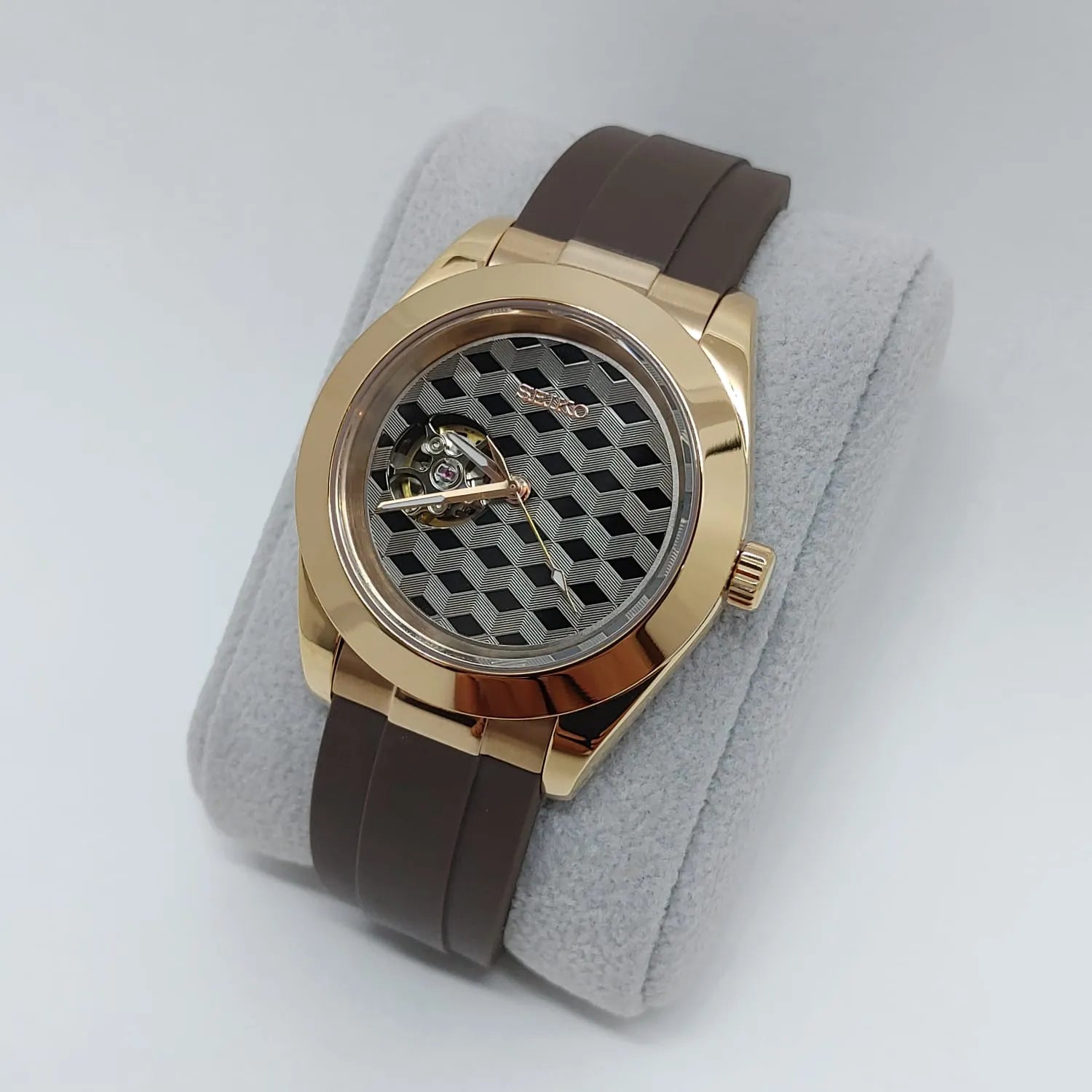 Gold-tone round watch with a grey hemicube guilloche dial and brown rubber strap.