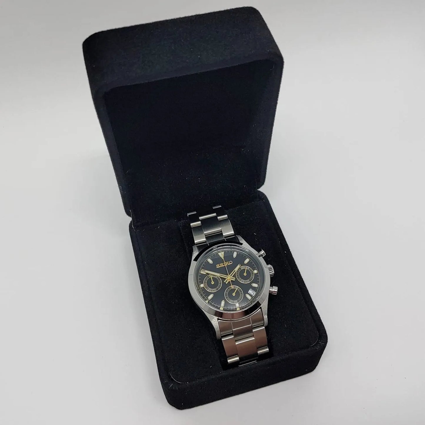 Seiko VK63 chronograph watch with black dial, gold-tone markers, and stainless steel bracelet in black velvet box.