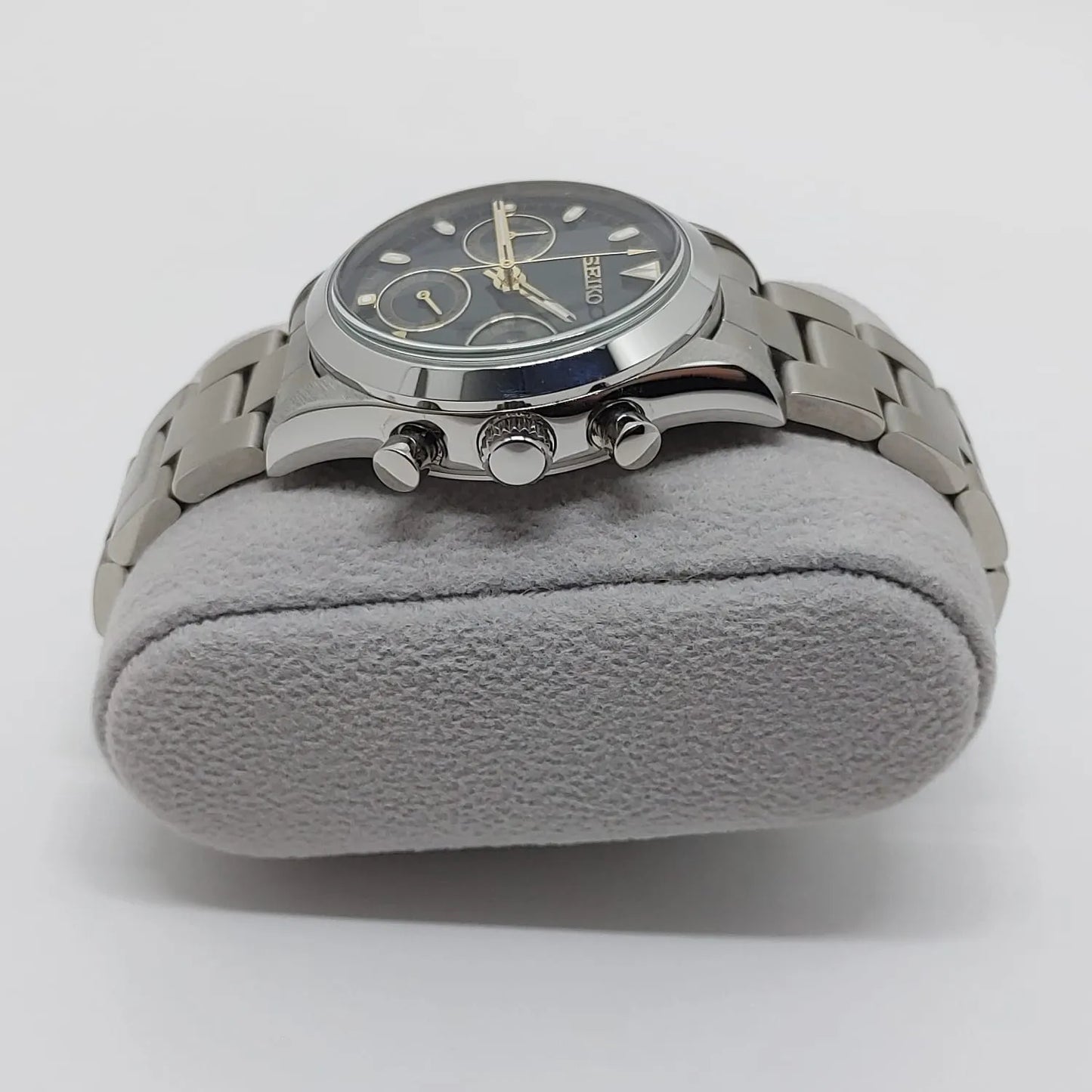 Stainless steel chronograph watch with black dial, cream subdials, and metal link bracelet on gray cushion.