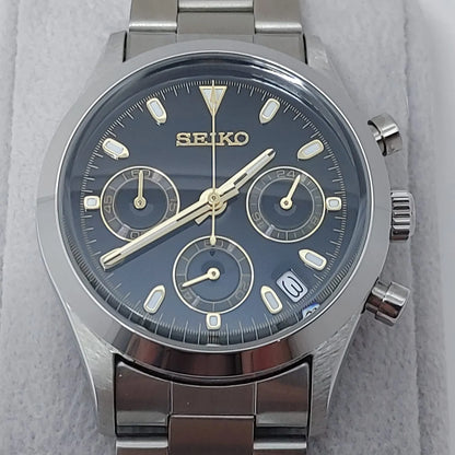 Seiko VK63 chronograph watch with black dial, gold-tone markers, three subdials, date window, and stainless steel bracelet.