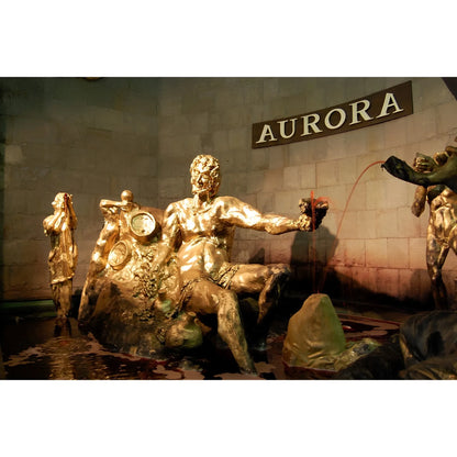 Bronze statue of a seated muscular man holding a small wine glass, with the word Aurora on a stone wall behind.
