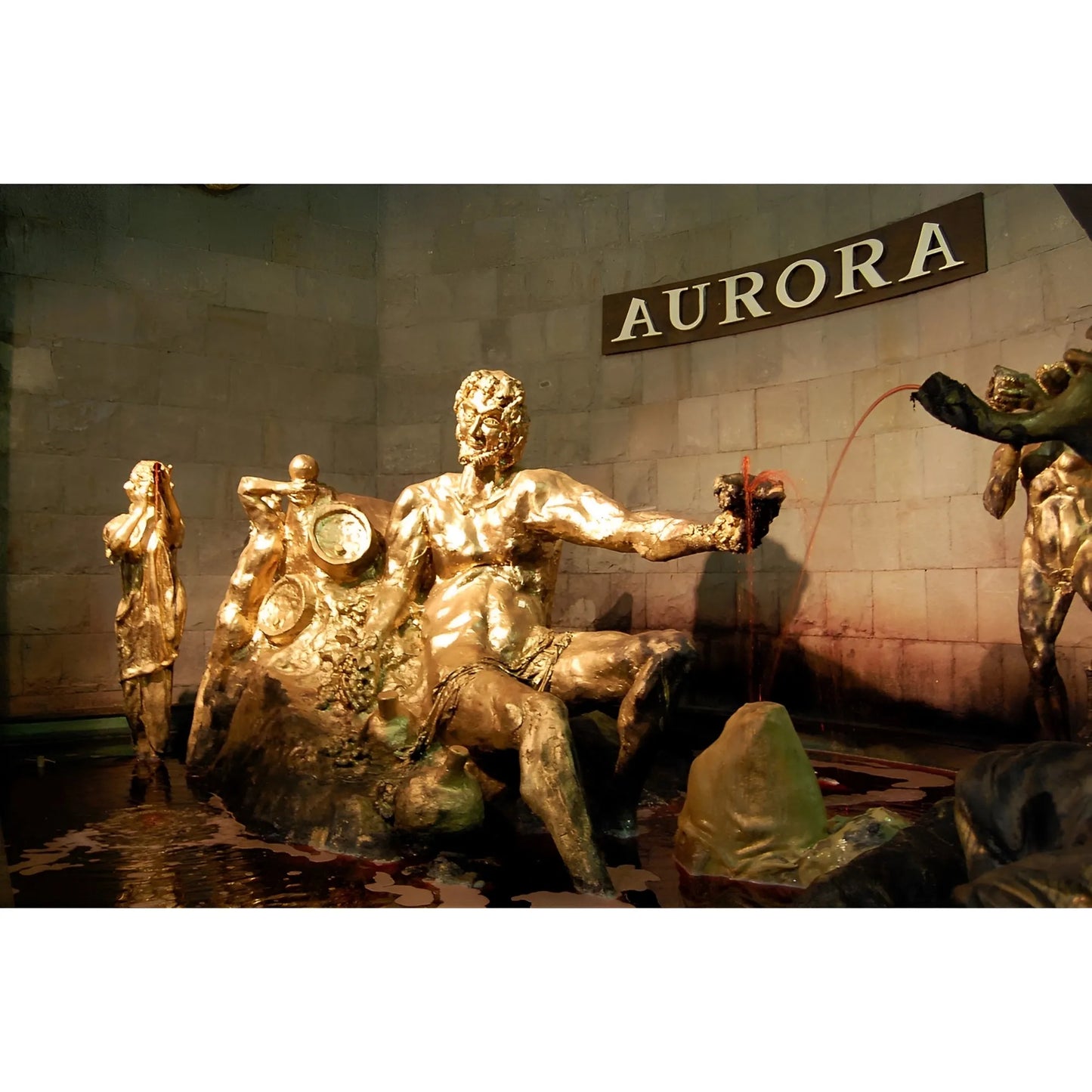 Golden bronze statue of a reclining man holding a wine glass, with a large urn and water feature, set against a stone wall.