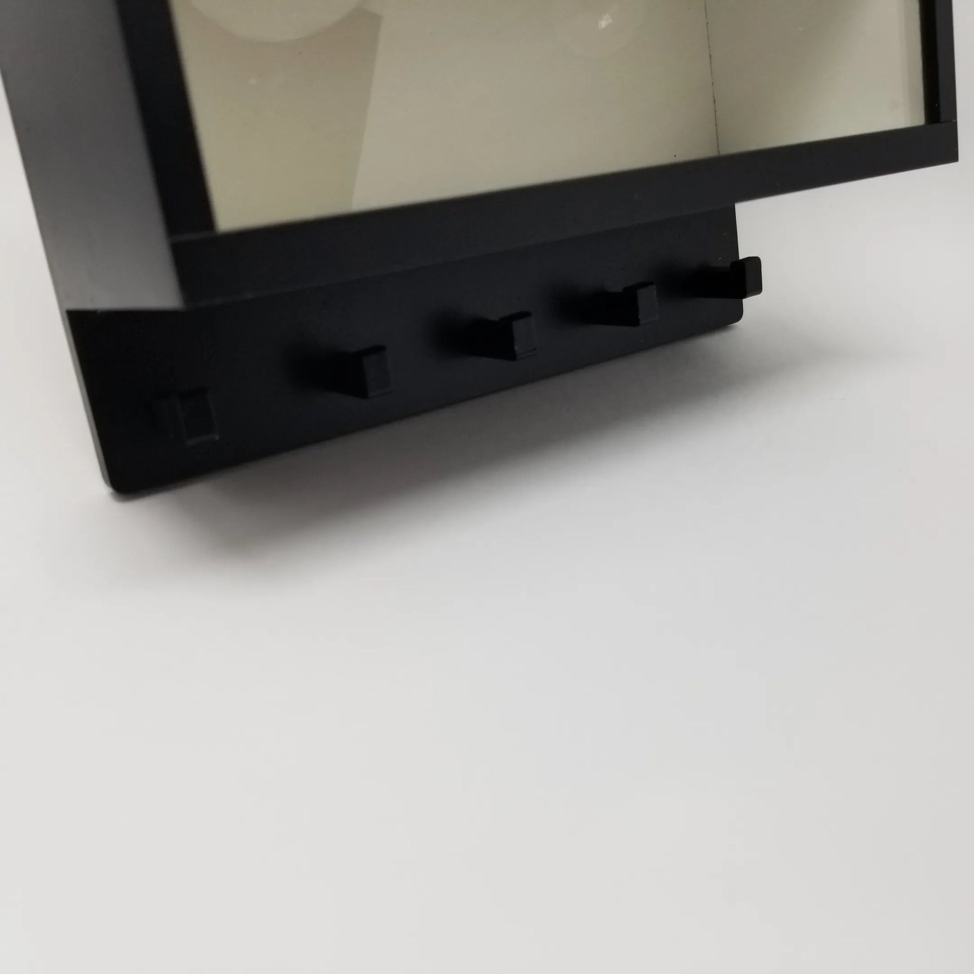 Black metal wall key hook rack with four evenly spaced hooks, attached to a clear display case frame.