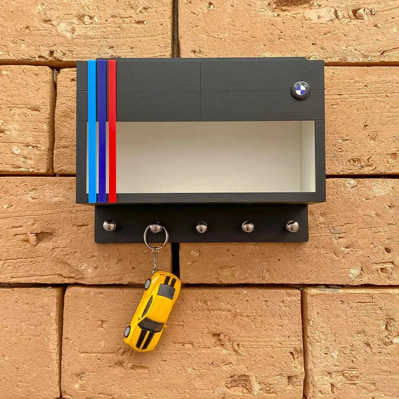 Black BMW wall key hook rack with clear display case, red and blue racing stripes, and five metal hooks holding a yellow.