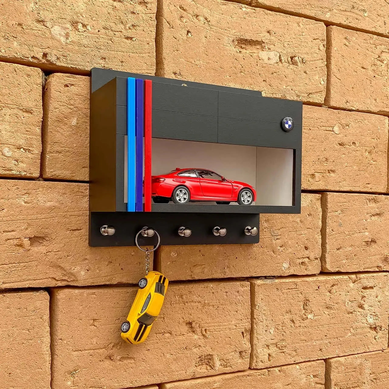 Black BMW wall-mounted key hook rack with red scale model car and four metal hooks on brick wall.