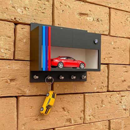 Black BMW wall-mounted key hook rack with red scale model car and four metal hooks on brick wall.