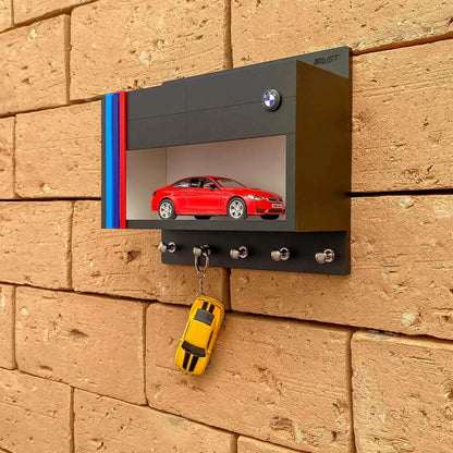 Black metal BMW wall key hook rack with red scale model car and five silver hooks mounted on brick wall.