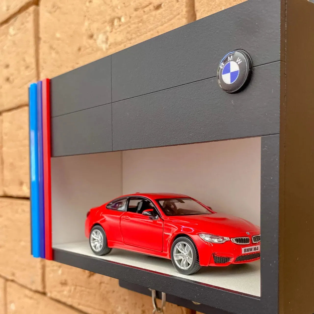 Black wall-mounted display case with horizontal slots, featuring a red scale model BMW car and BMW logo badge on the upper.