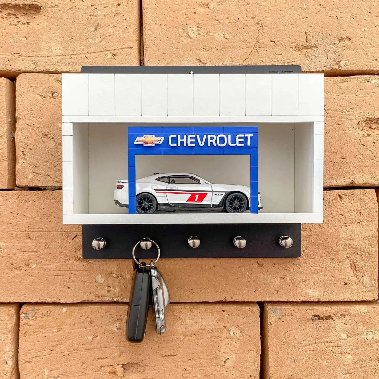 Black and white Chevrolet wall key hook rack with silver hooks and a scale model car inside a display case.