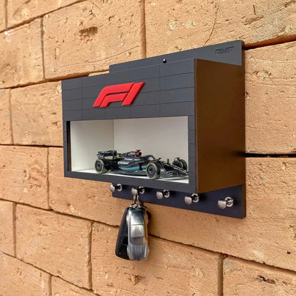 Black rectangular wall-mounted key hook rack with a red F1 logo and a clear display case holding a small Formula One car.