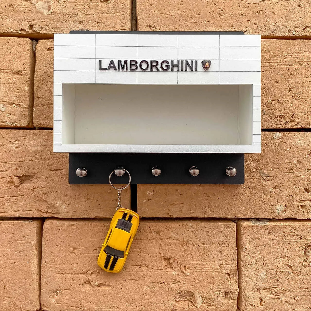 White and black wall-mounted key hook rack with Lamborghini logo and five metal hooks, holding a yellow car-shaped keychain.