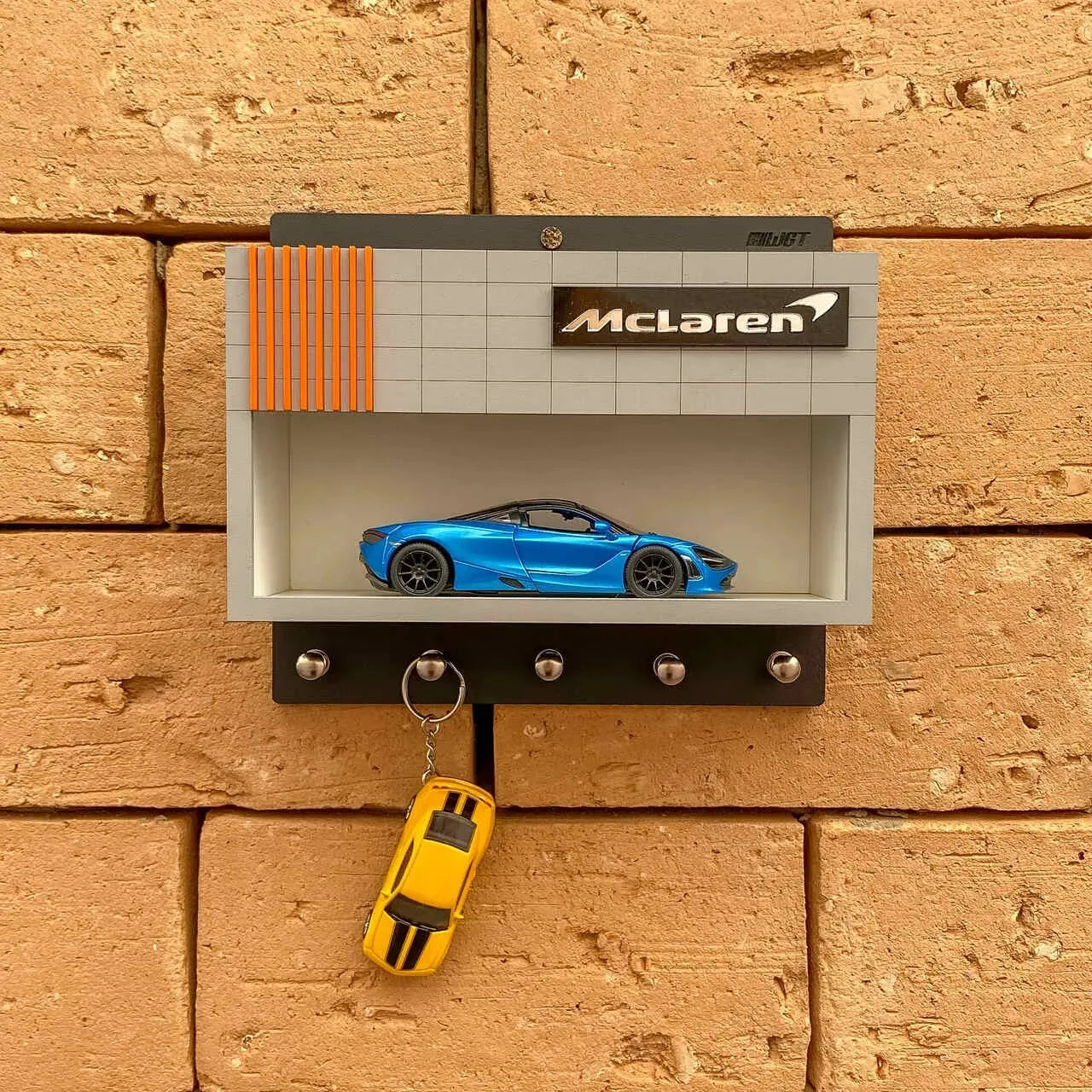 Black wall-mounted key hook rack with a gray shelf, orange stripe detail, McLaren logo, and blue scale model car on display.