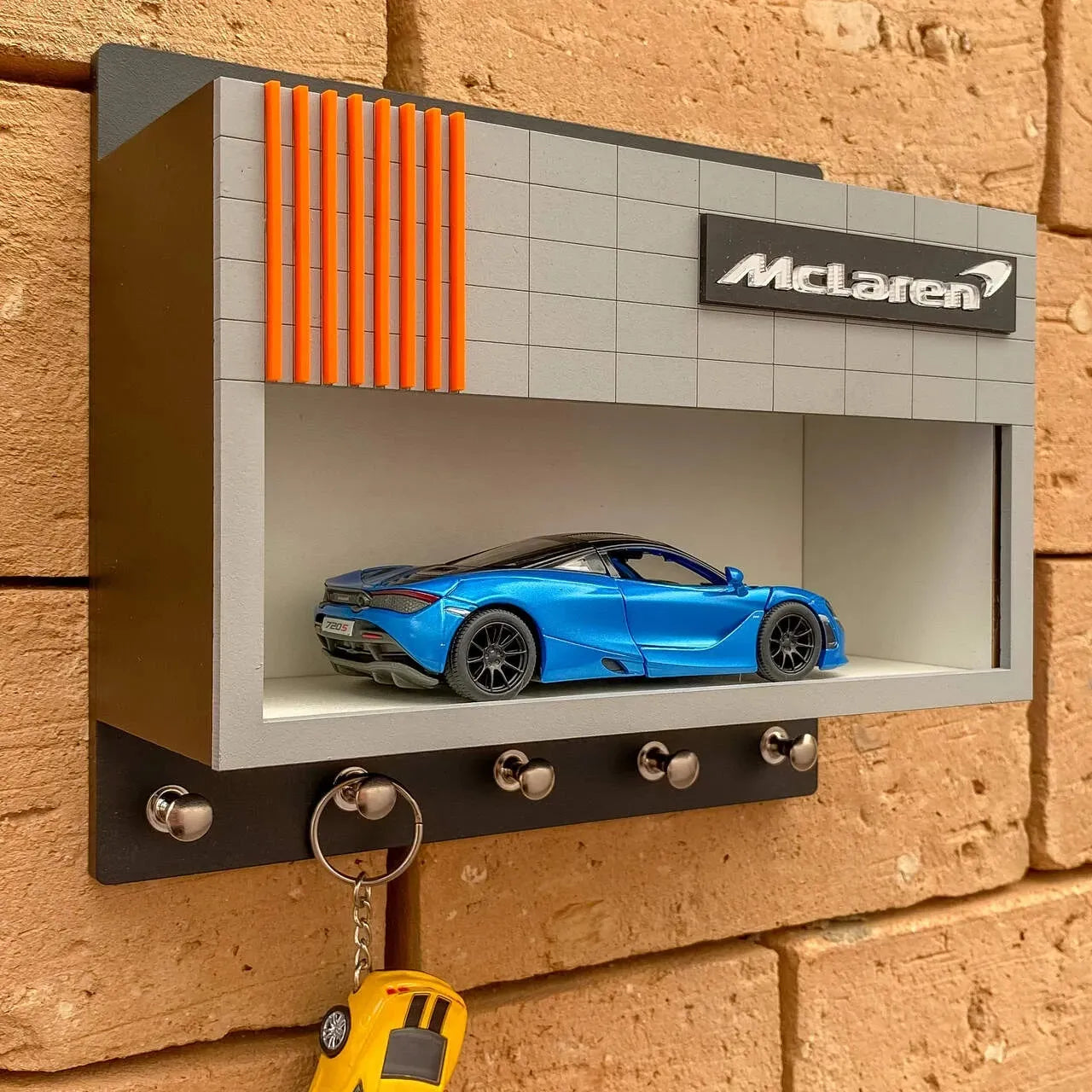 Gray McLaren key hook rack with orange stripes and blue scale model car display on a brick wall.