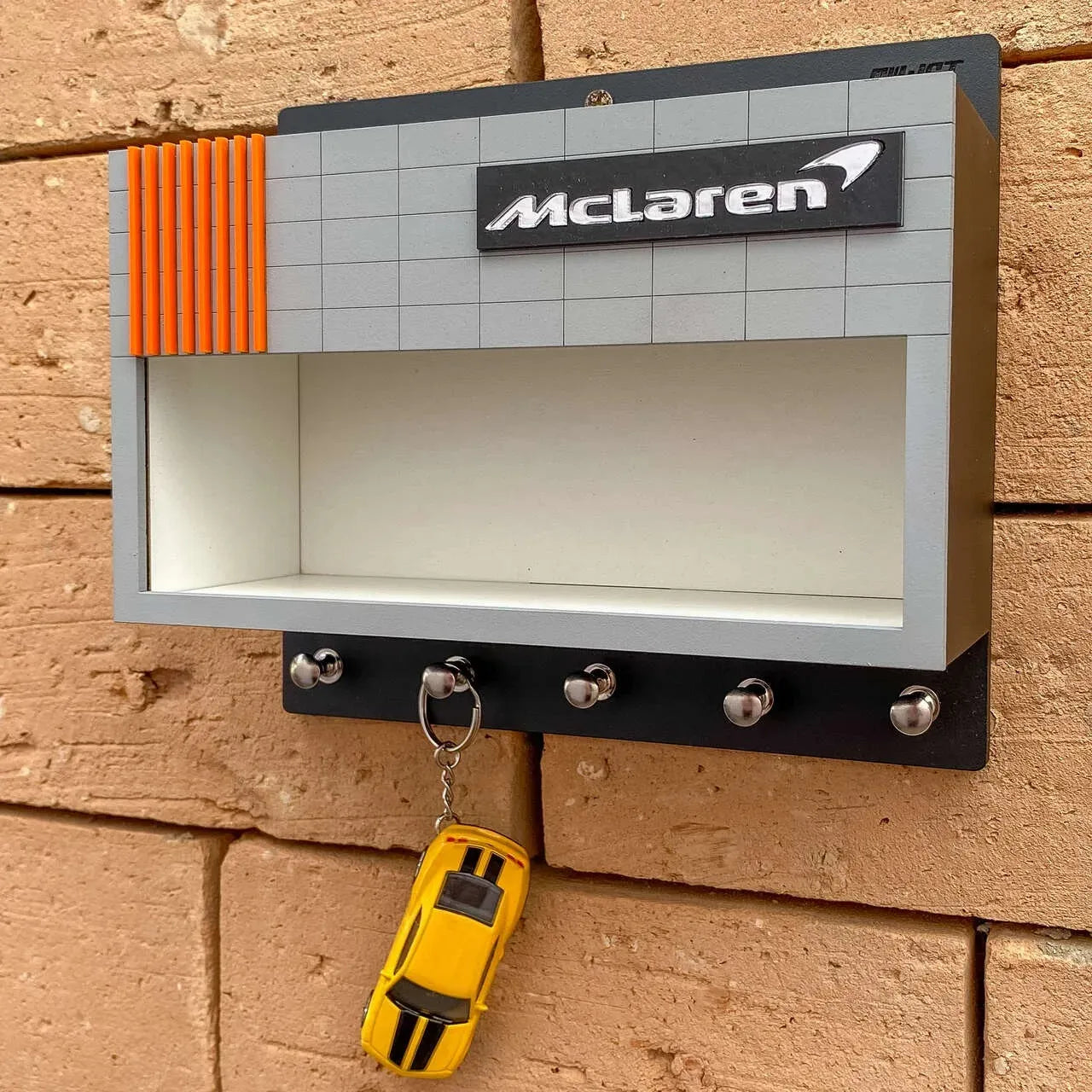 Gray rectangular wall-mounted McLaren key hook rack with orange accent and four metal hooks, one holding a yellow toy car.