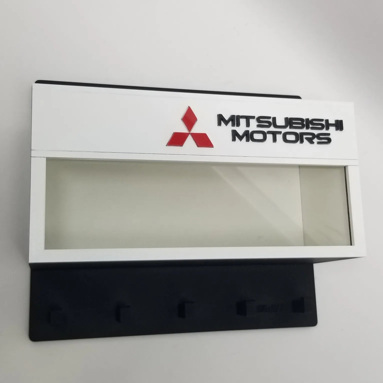 White rectangular Mitsubishi Motors wall key hook rack with clear display case and four black hooks.