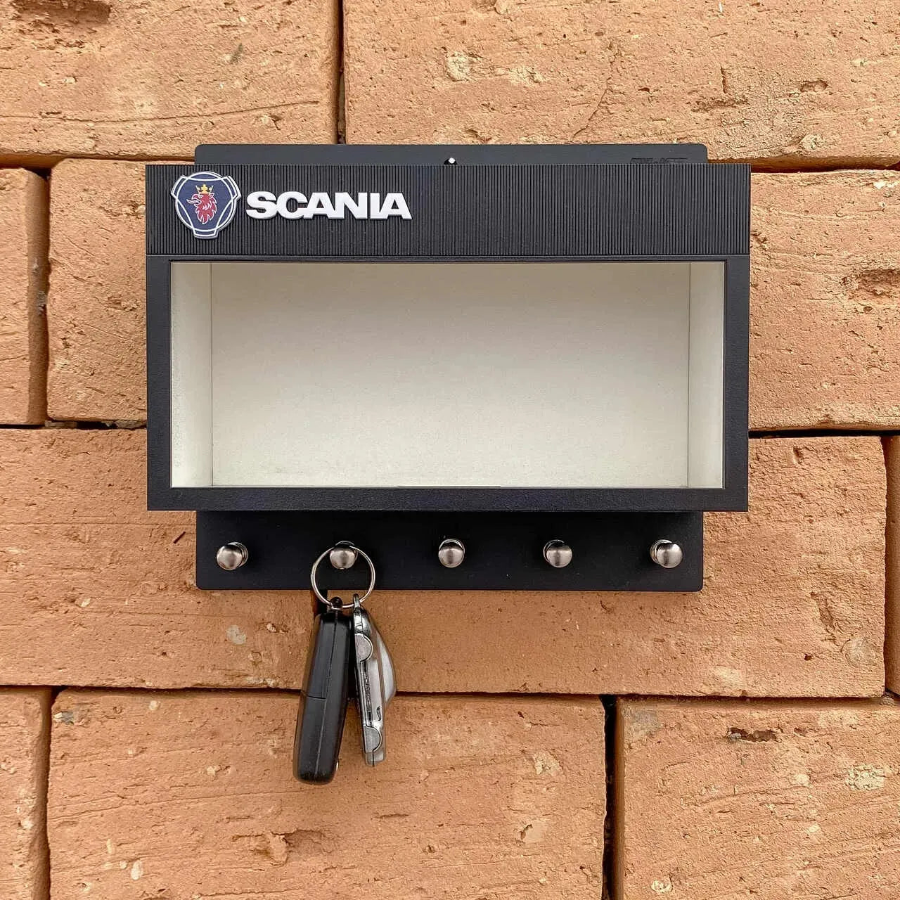 Black rectangular Scania wall key hook rack with four metal hooks and a clear display case on a brick wall.