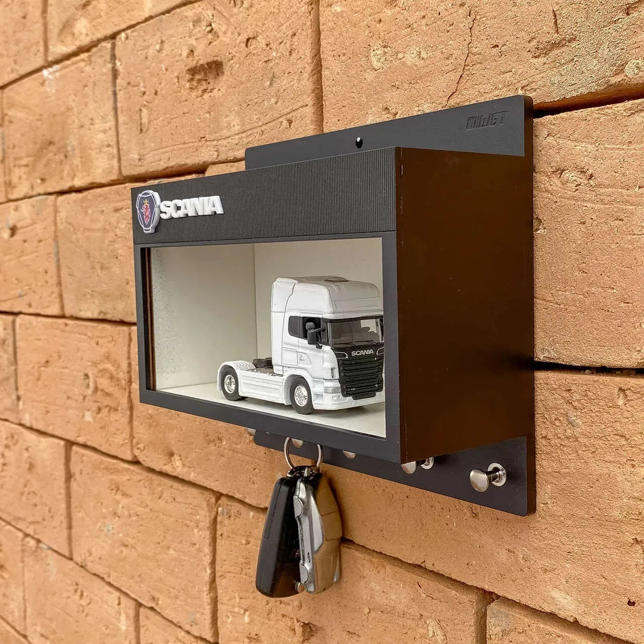 Black metal wall-mounted key hook rack with a glass display case featuring a white scale model truck inside and two keys.