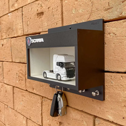 Black metal wall-mounted key hook rack with a glass display case featuring a white scale model truck inside and two keys.