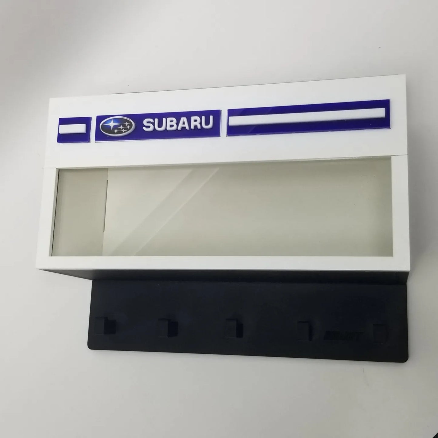White rectangular Subaru wall key hook rack with glass display case and four black hooks.